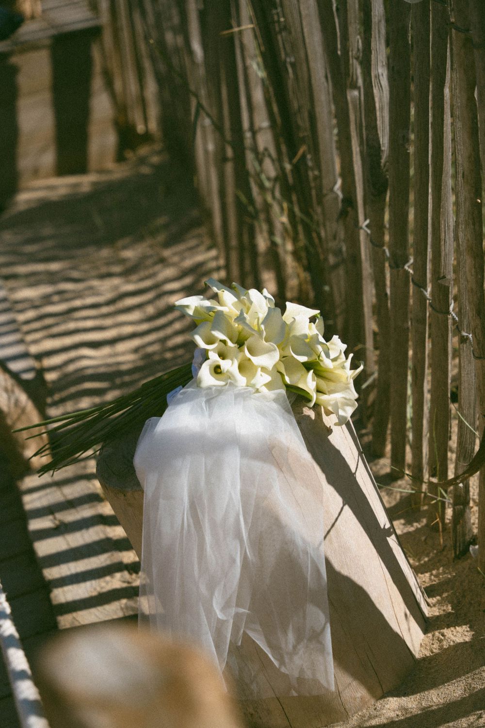 A white bridal veil with calla lily flowers rests against a wooden fence casting dramatic shadows.