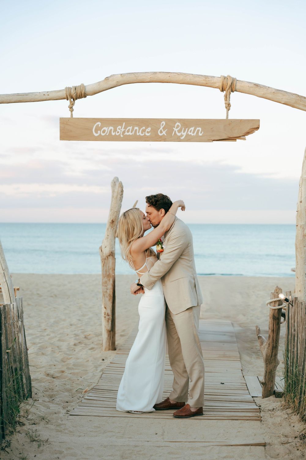 Un couple partage un baiser romantique sur une promenade au bord de la plage sous une arche en bois avec un signe nominative au coucher du s