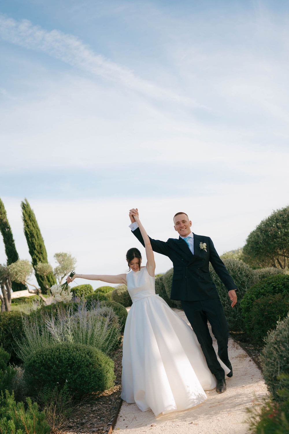 Joyful newlyweds raise hands in celebration on a garden path surrounded by lavender and cypress trees under a blue sky.