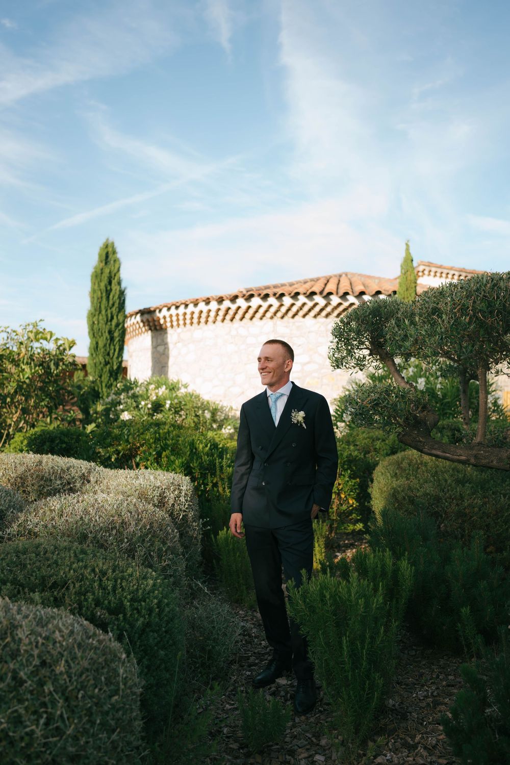 Groom in black suit with boutonniere stands among lavender bushes outside a stone villa under blue sky.