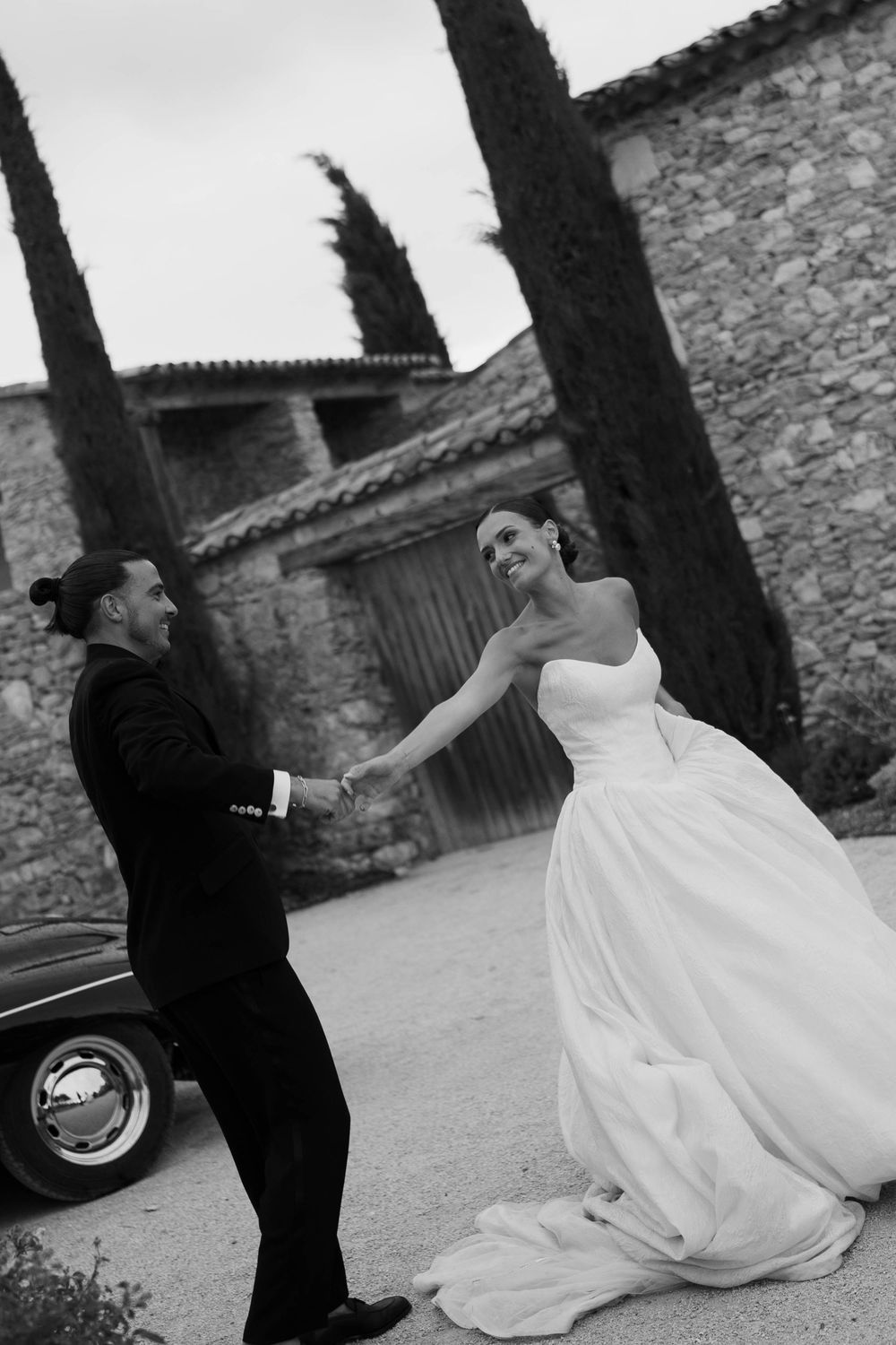 Black and white photo of a bride and groom holding hands, smiling near a vintage car with a stone building backdrop.