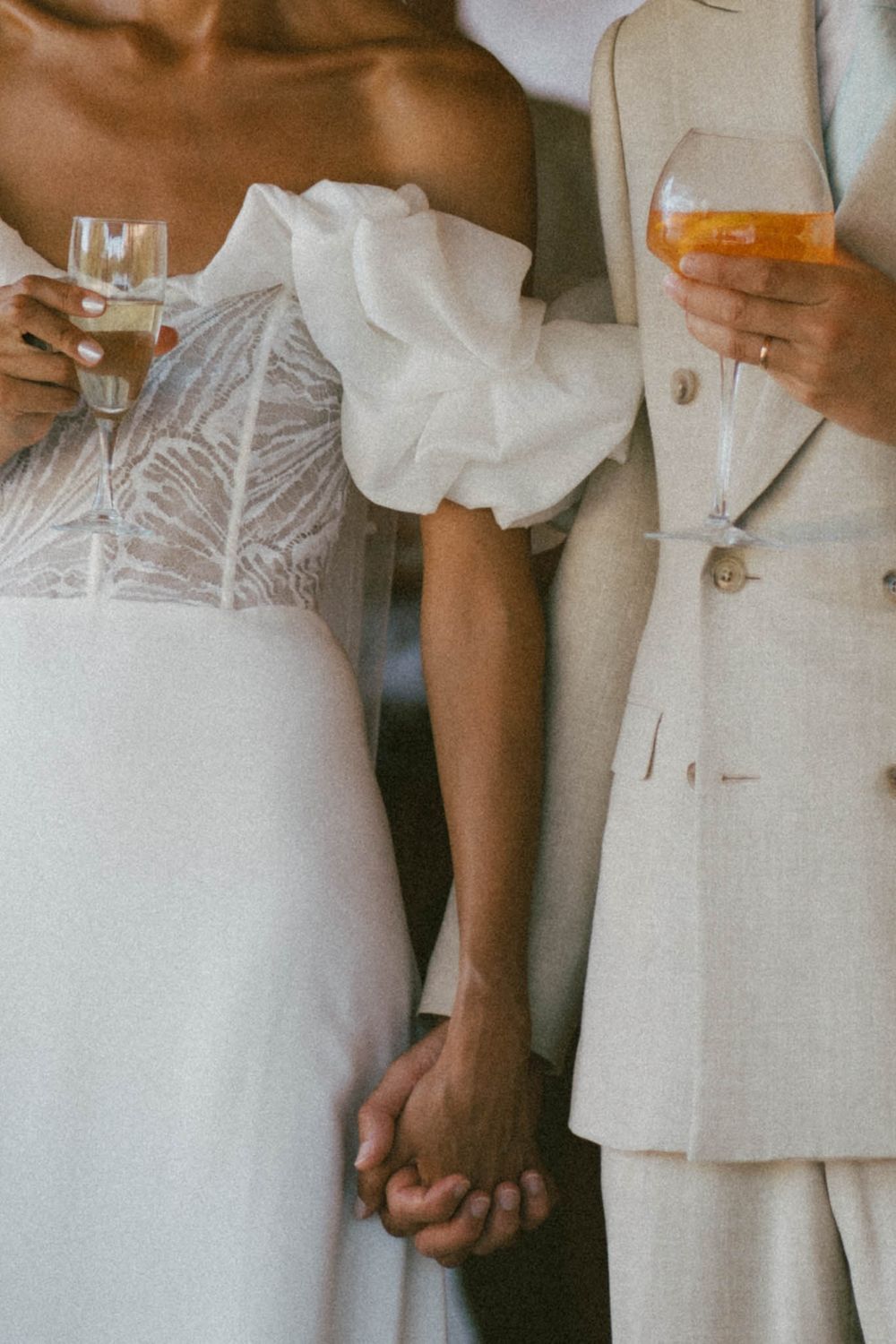 Two brides holding hands in elegant white wedding attire, each holding a champagne glass at their wedding celebration.