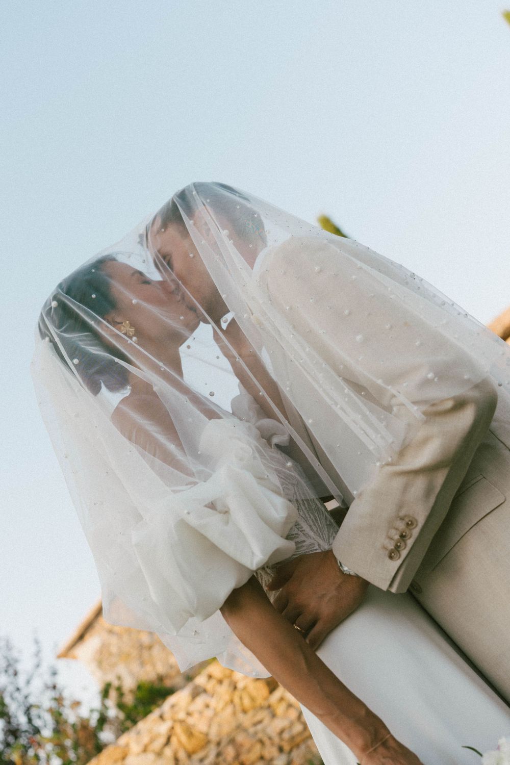 Bride and groom sharing a romantic kiss under a pearl-adorned veil against a clear blue sky.