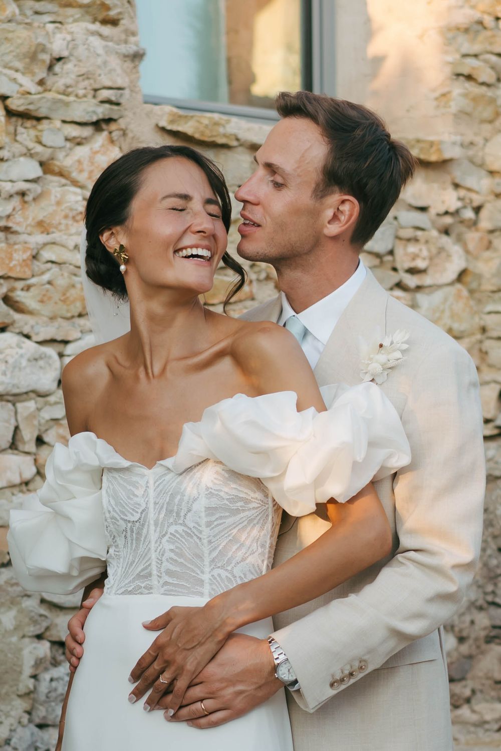 La mariée et le marié s'embrassent joyeusement devant un mur de pierre, elle porte une robe blanche à épaules dénudées, il porte un costume