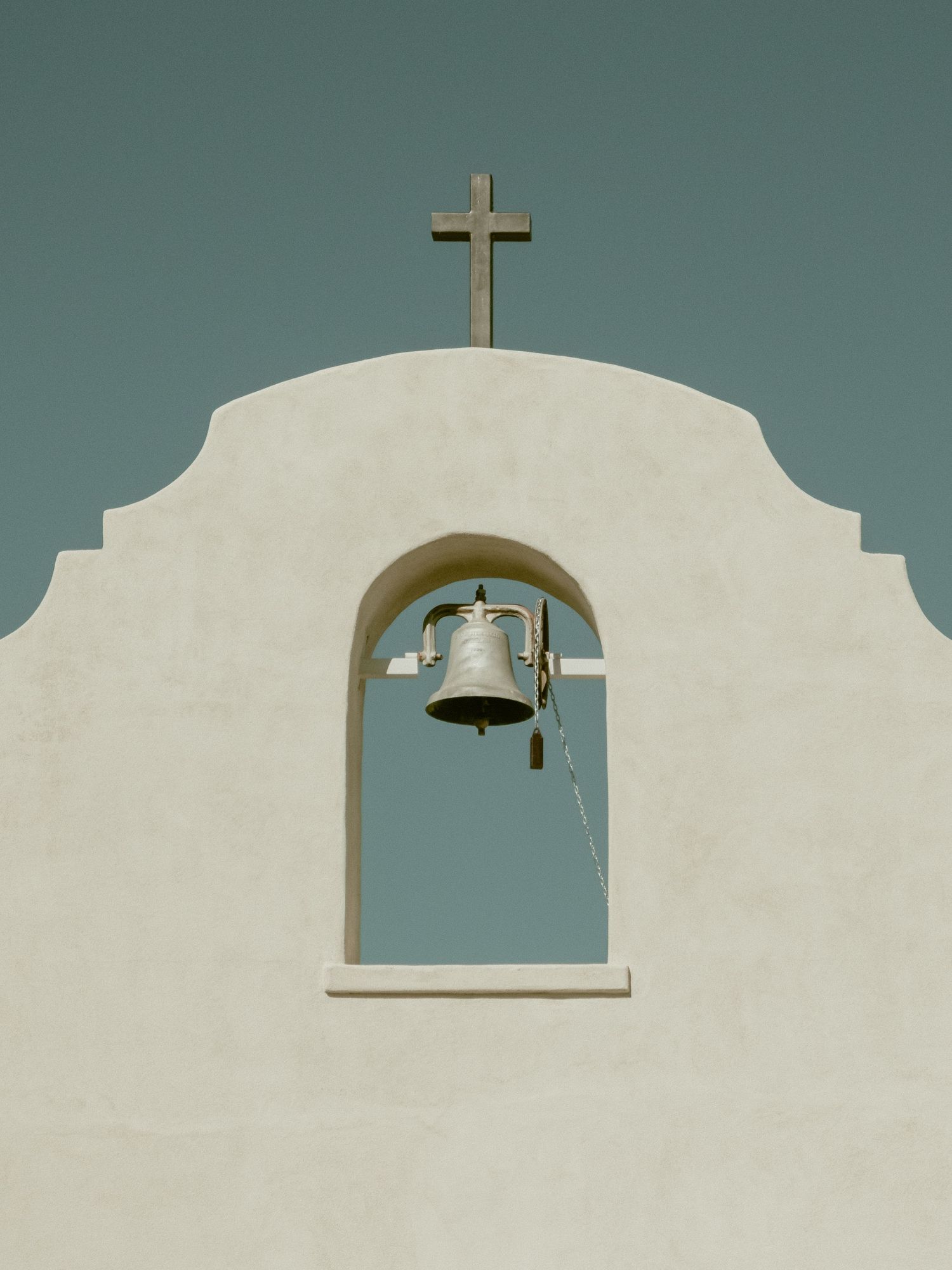 Minimalist white church bell tower with cross and bell against teal sky.