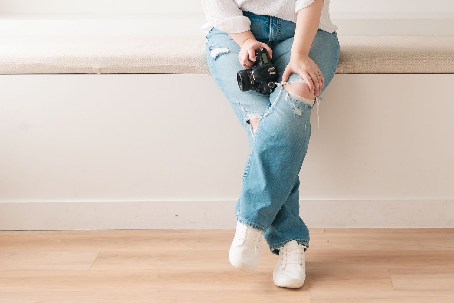 A person in light blue distressed jeans and white sneakers sits on a wooden floor while holding a camera.
