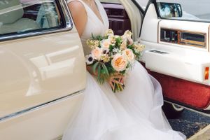 A peach and white rose wedding bouquet rests on a flowing white tulle dress in a vintage car with cream interior.