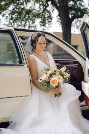 A beautiful white wedding dress with flowing tulle cascades from a vintage cream-colored car as a bouquet of peach roses is held.