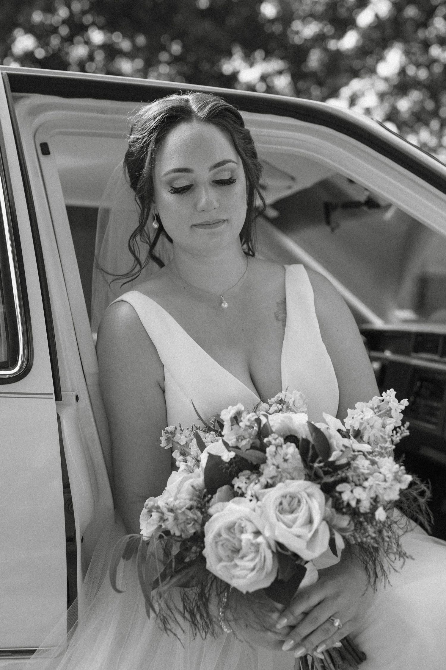A bride in a white wedding dress holds a beautiful bouquet of roses and flowers while sitting in a vintage car.
