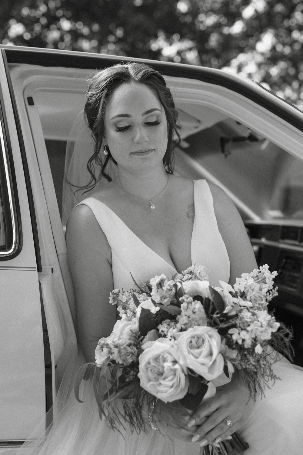 A bride in a white wedding dress holds a beautiful bouquet of roses and flowers while sitting in a vintage car.