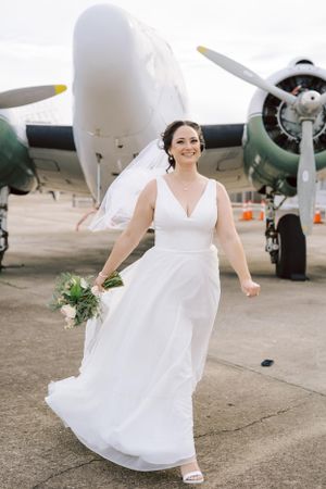 A bride in a flowing white wedding dress holds a bouquet while standing in front of a vintage aircraft with propellers.