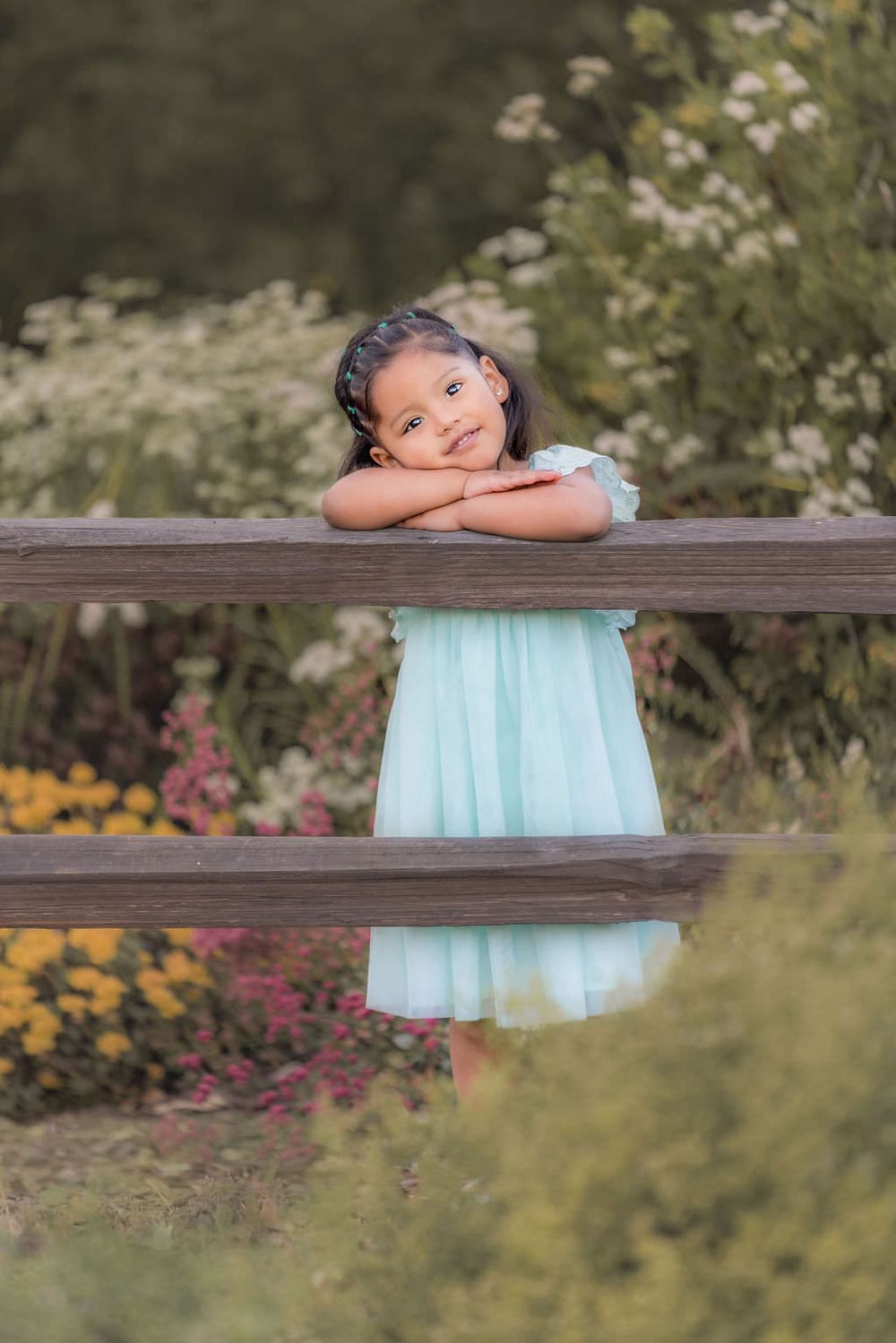 Child in turquoise dress leaning on wooden fence surrounded by wildflowers.
