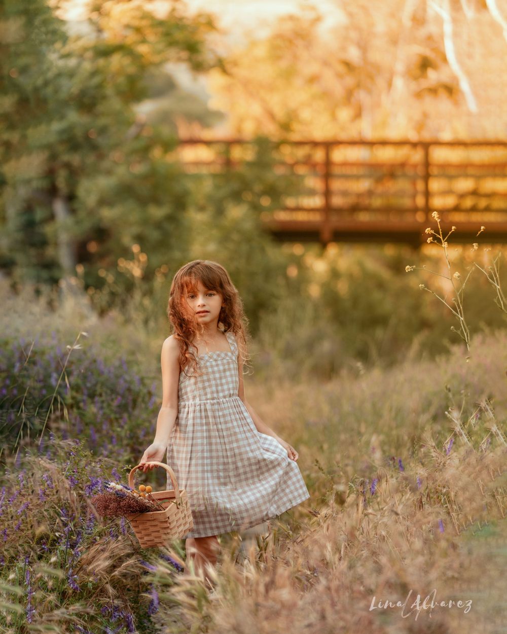 A gingham dress-clad figure holds a wicker basket near purple wildflowers and a wooden bridge at golden hour.