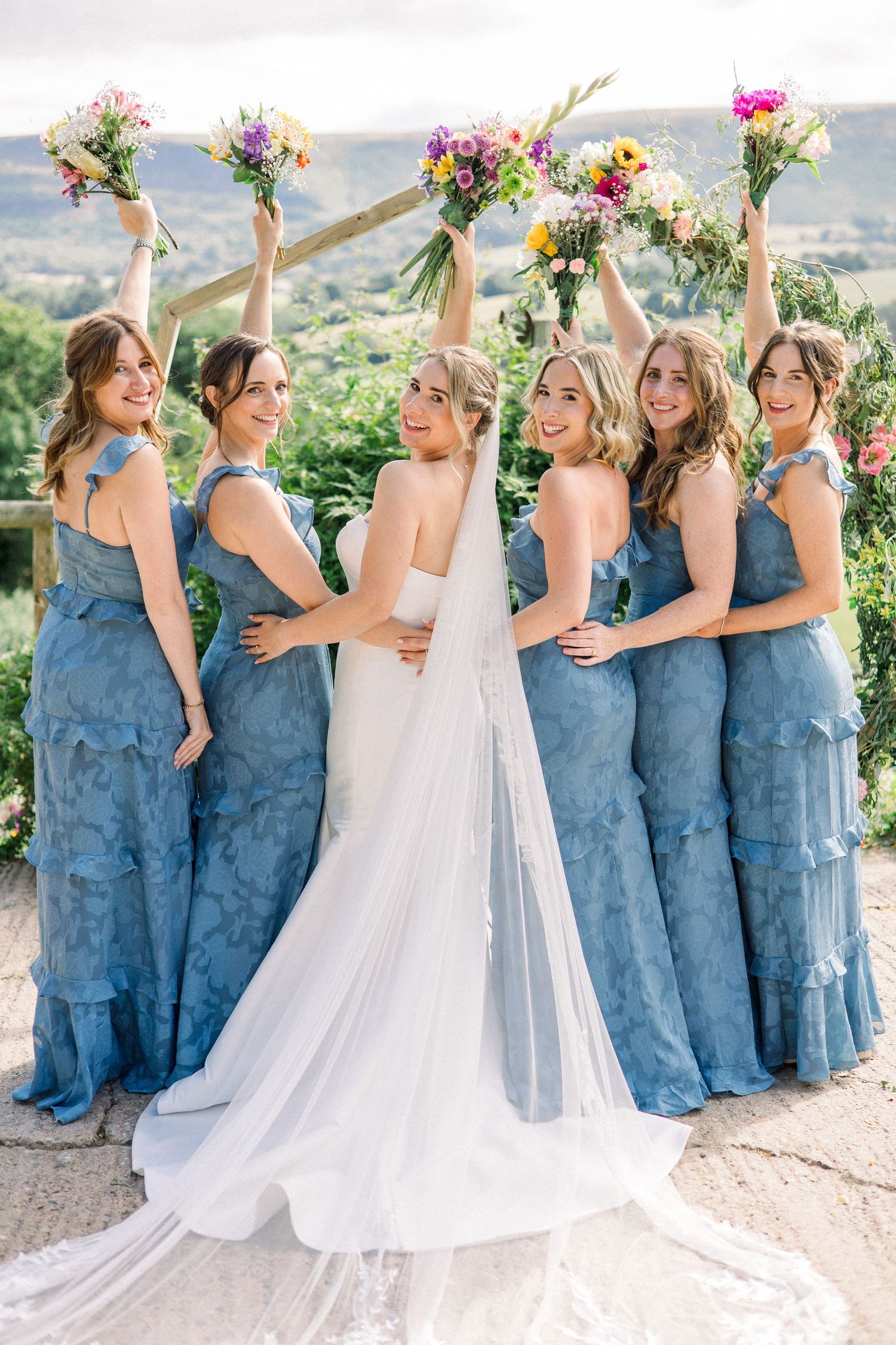 Bridesmaids in blue dresses celebrate with bride against mountain backdrop.