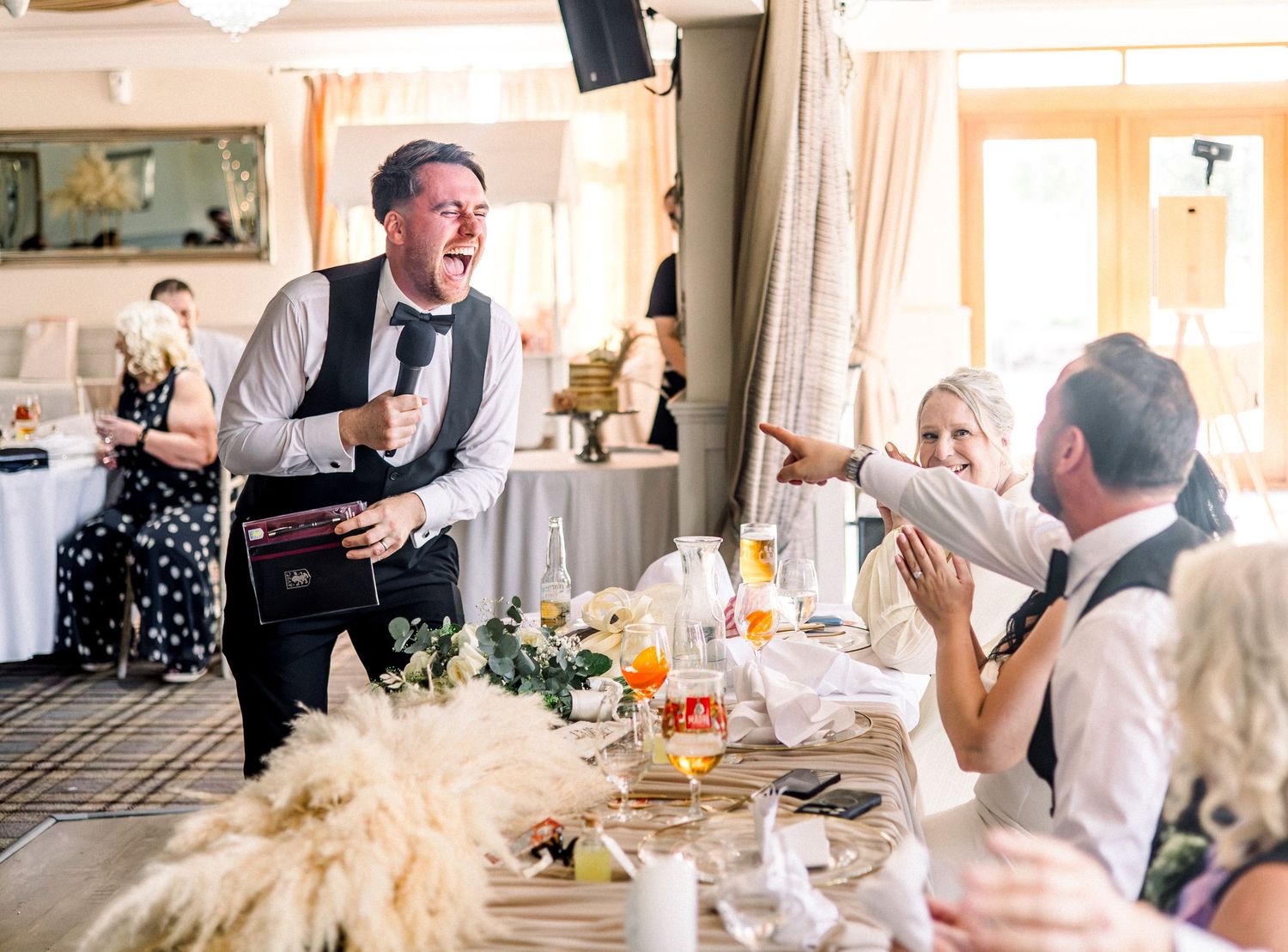 Energetic wedding MC performs speech at elegant reception, engaging laughing guests at decorated table.