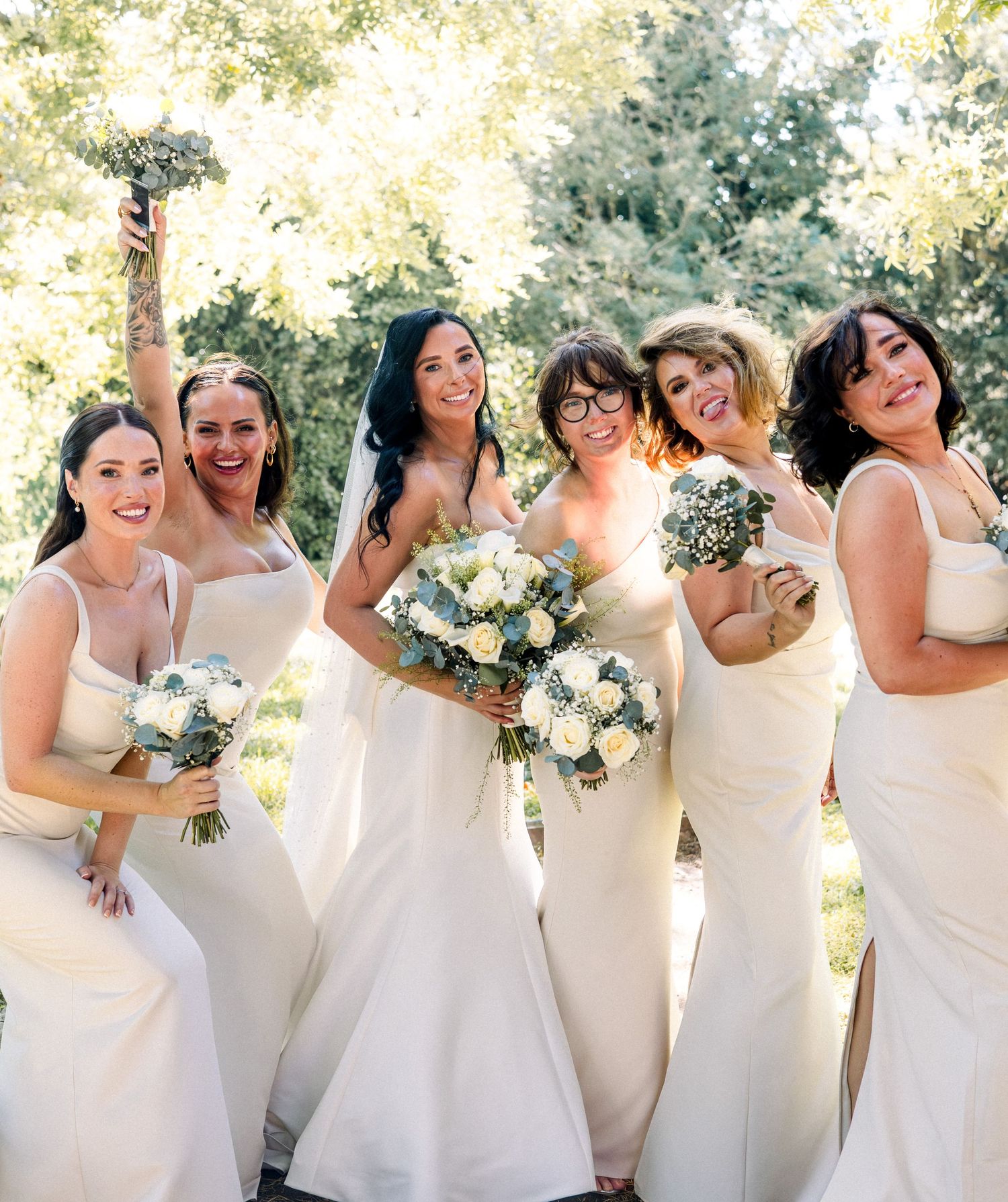 Bride and bridesmaids in ivory gowns laughing and posing outdoors with white floral bouquets on wedding day.