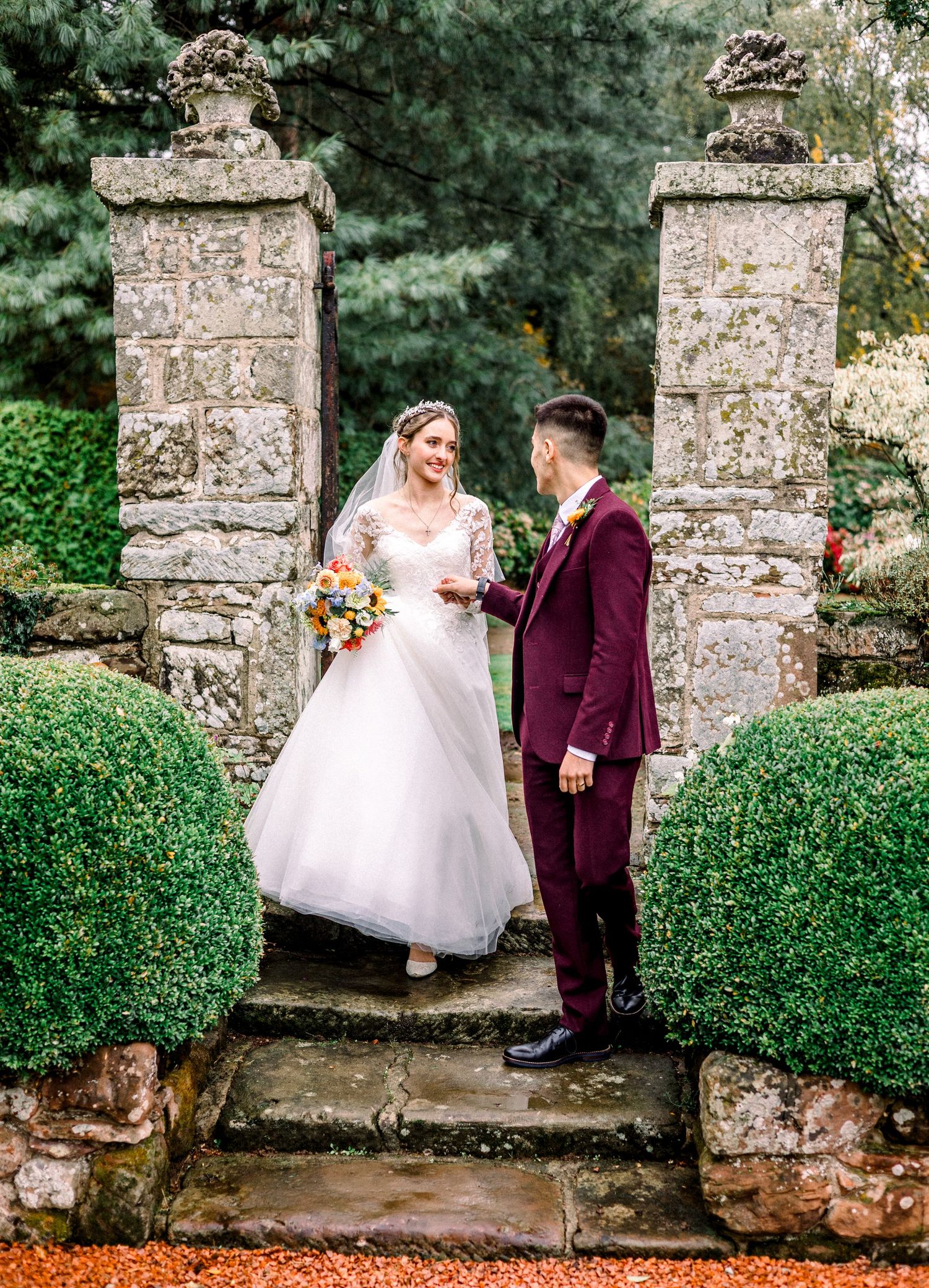 Wedding couple posing between stone pillars and manicured hedges in a formal garden setting.