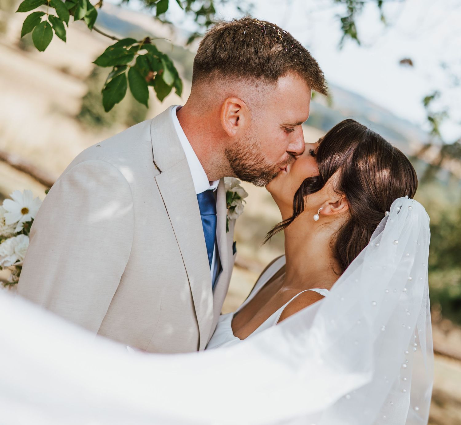 A bride and groom share a romantic kiss outdoors, with the bride's veil flowing in the breeze on their wedding day.