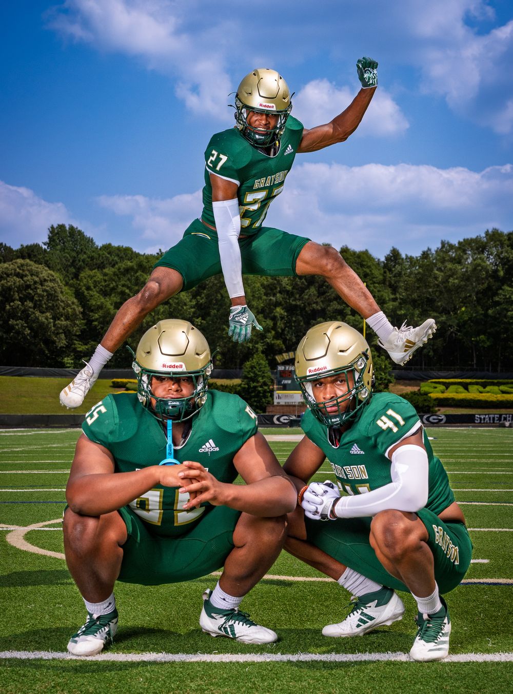 Three football players in green and gold uniforms pose for a creative team photo on a sunny field.