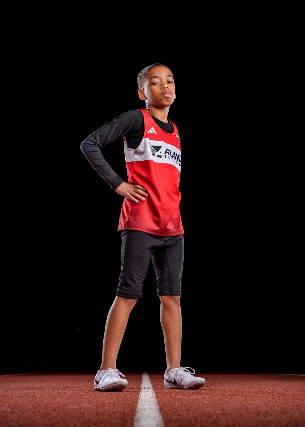 A young athlete stands confidently in a red and black track uniform on a running track against a dark background.