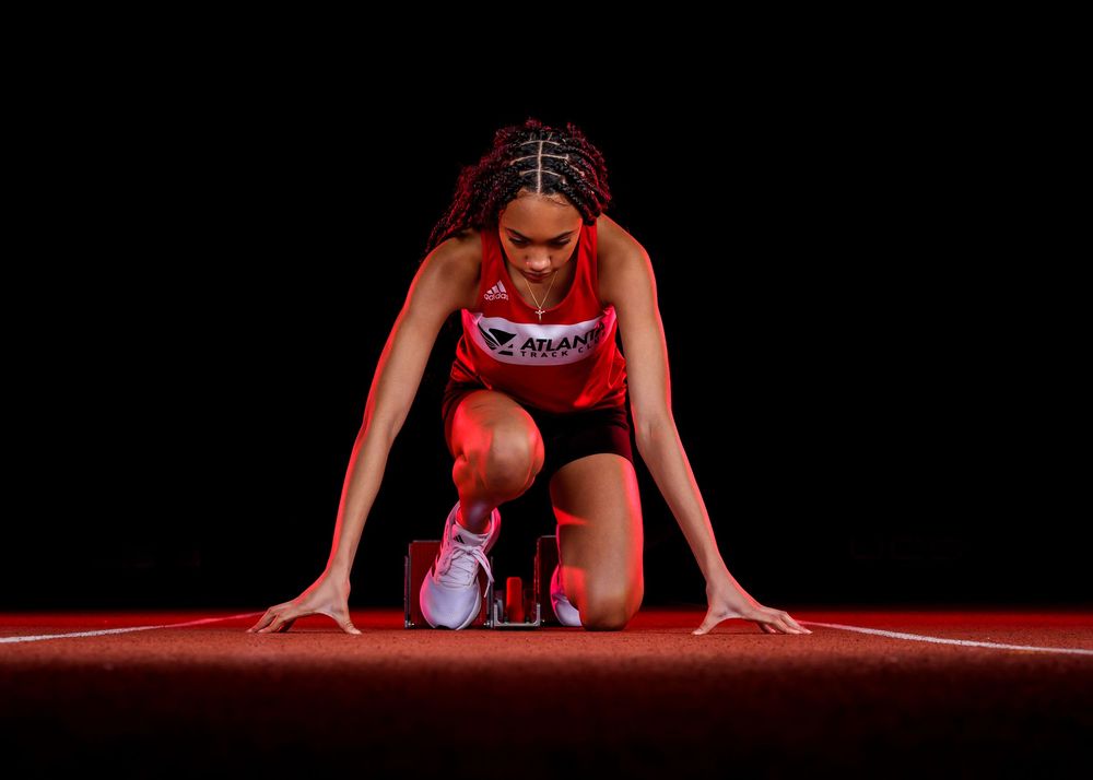 Athlete in red uniform crouching at starting position on track in dramatic lighting against dark background.