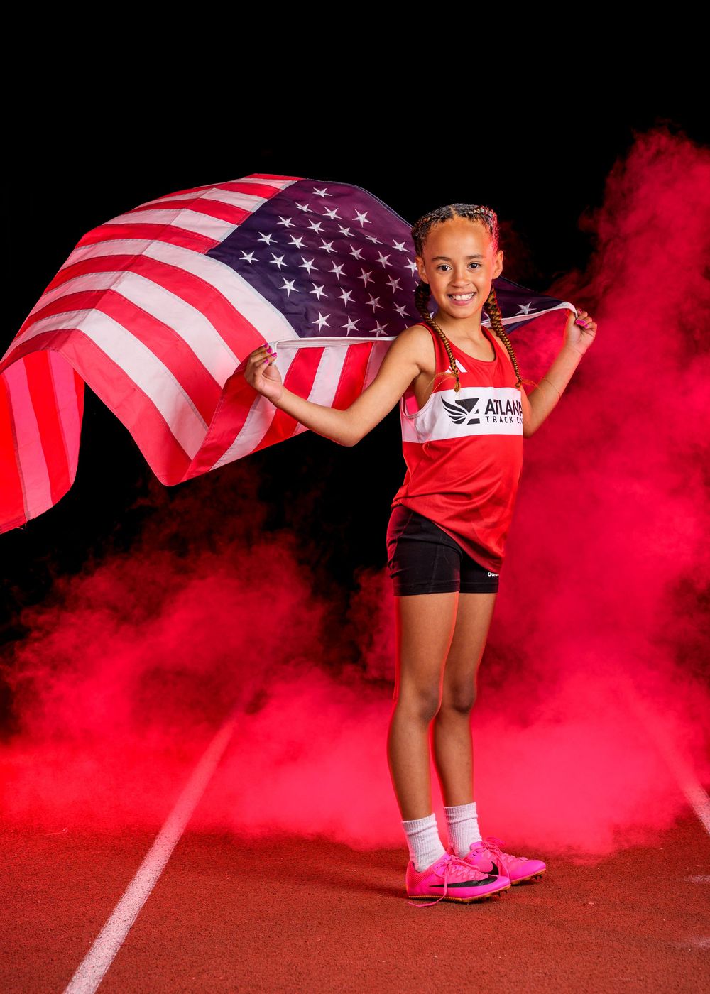 Track athlete poses with American flag amidst dramatic red smoke on running track wearing Team USA uniform.