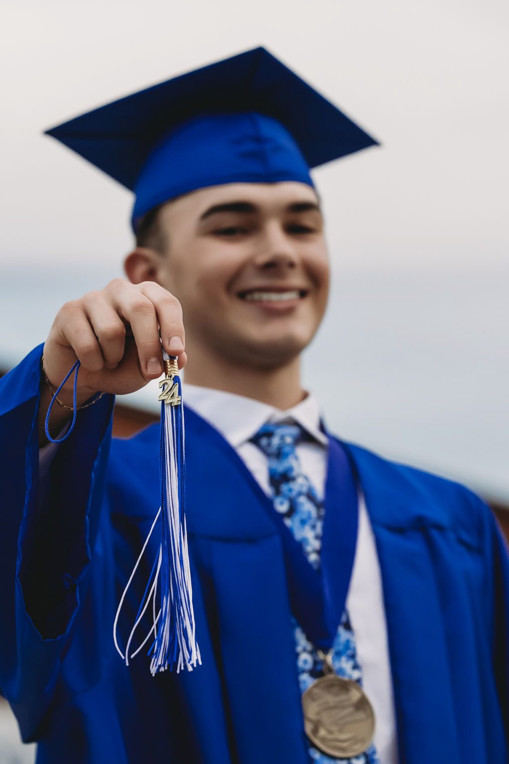 Graduate in blue cap and gown holds up graduation tassels while smiling at commencement ceremony.