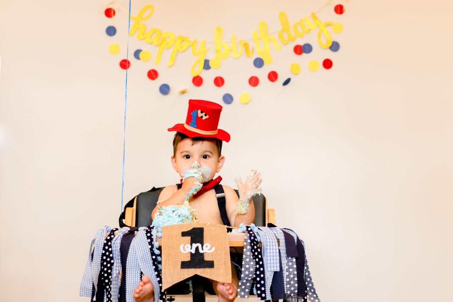 Baby in first birthday hat and decorative highchair sitting in front of colorful birthday party backdrop.