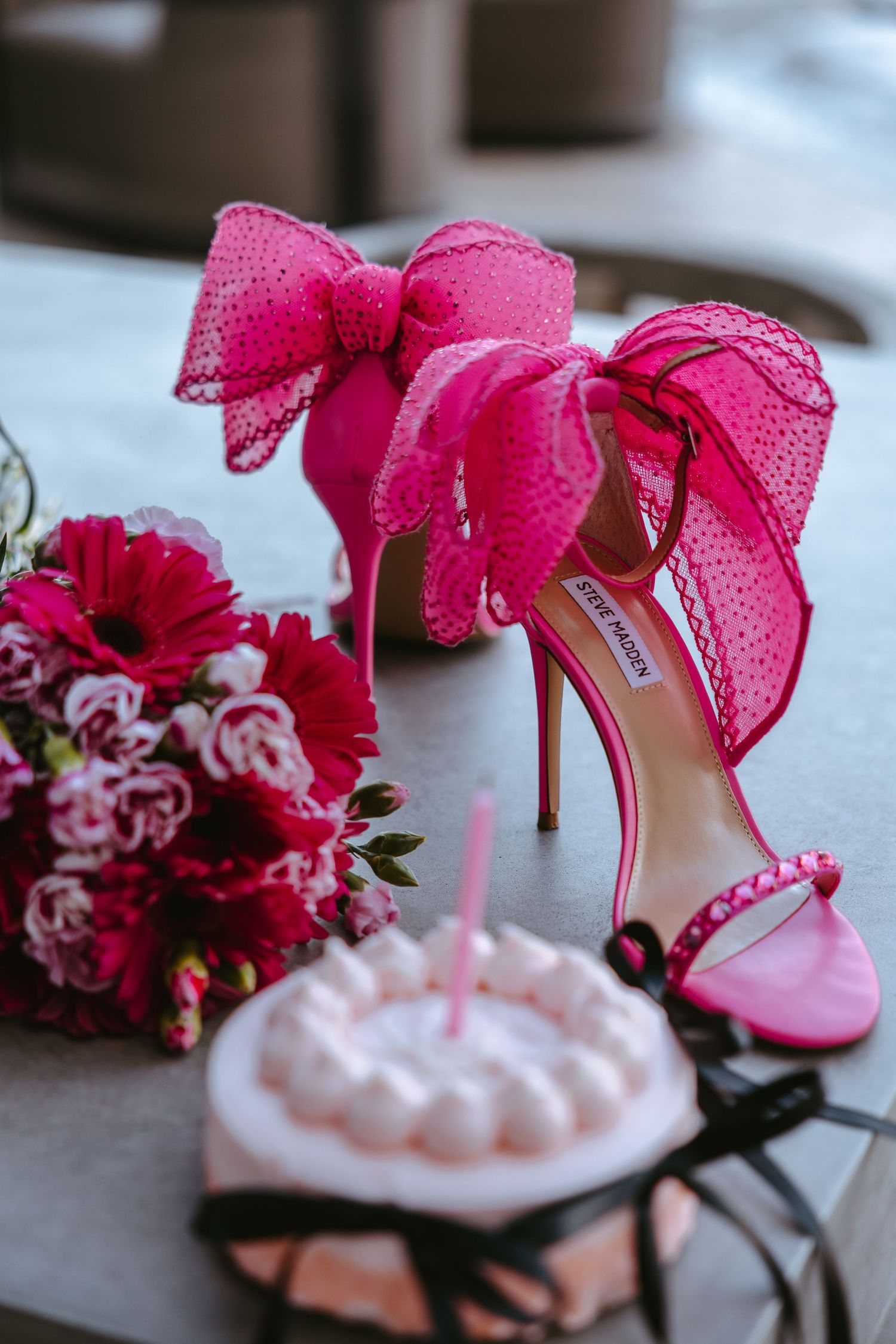 Pink bow heels next to floral bouquet and birthday cupcake on table.