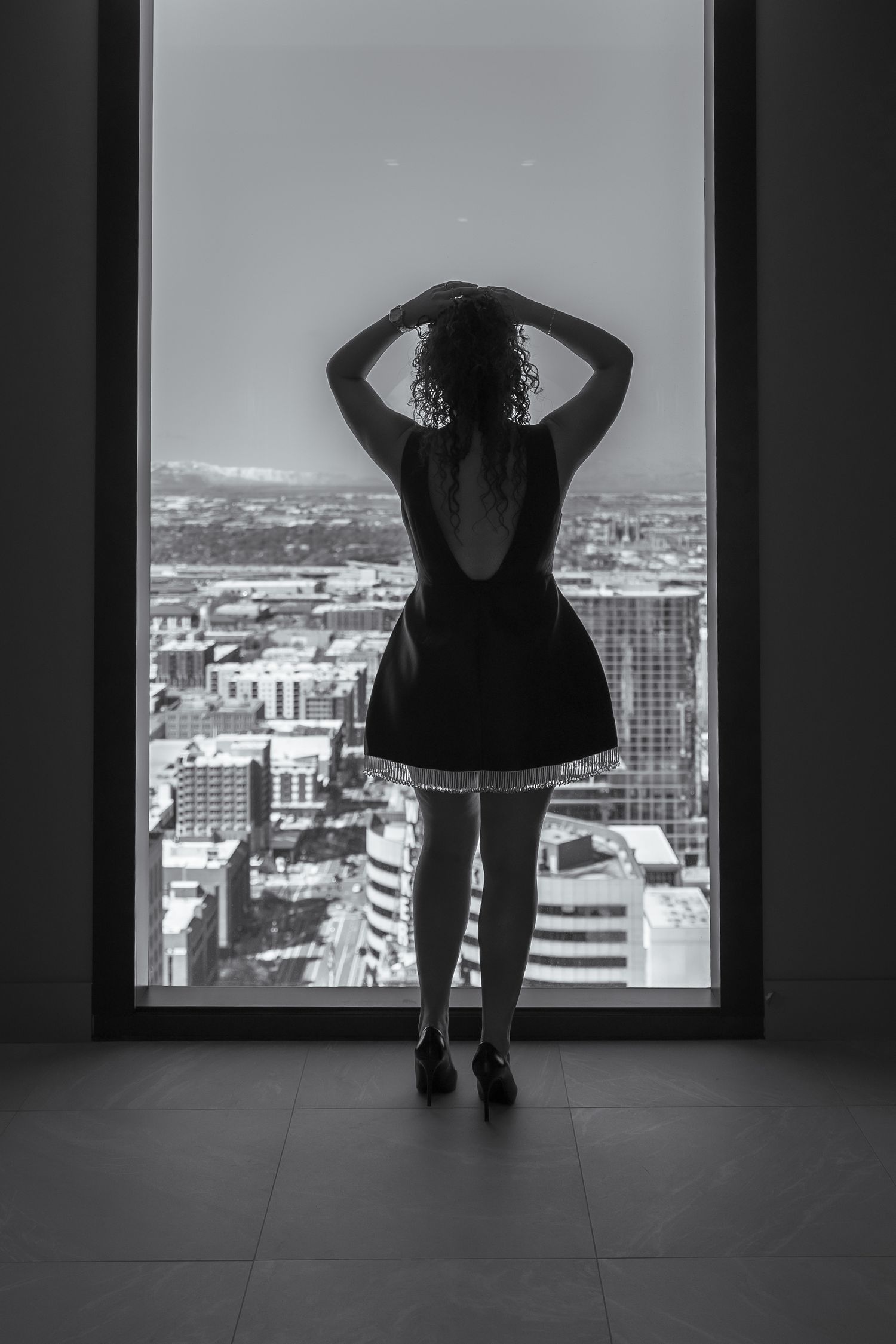 Silhouette of person in dress looking out large window at city view.