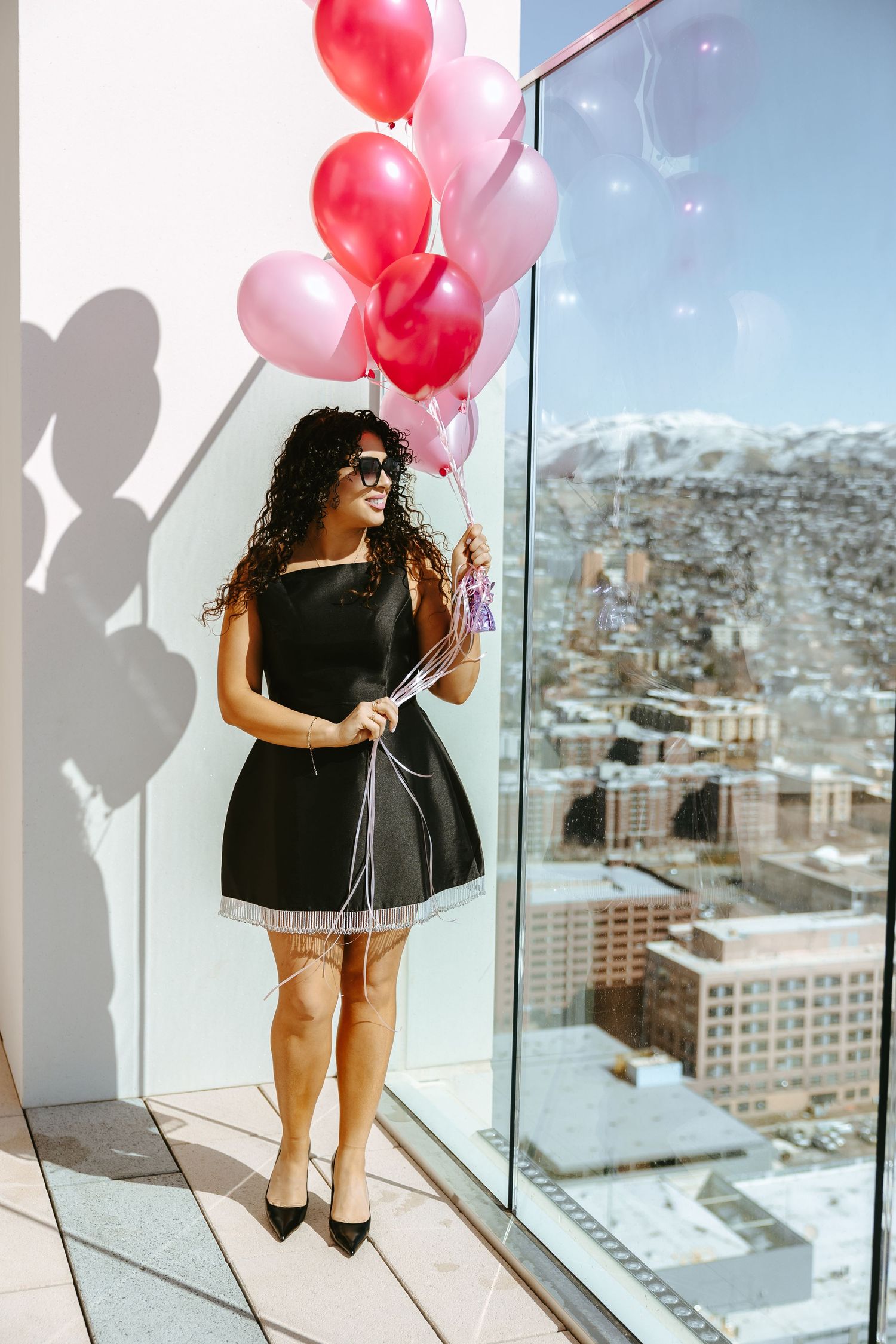 Woman in black dress holds pink balloons on high-rise balcony overlooking city.