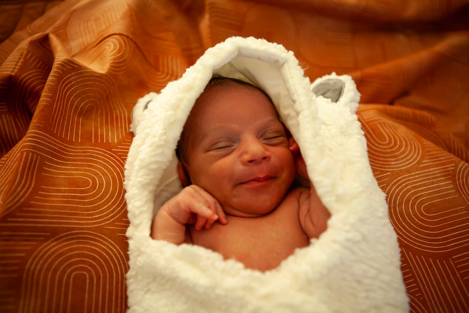 A newborn wrapped in a soft white blanket smiles peacefully against an orange textured background.