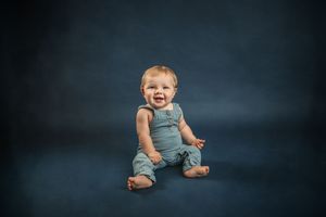 A happy baby in a blue romper sits barefoot on a dark blue studio background, smiling joyfully for marietta fine art children's photographer