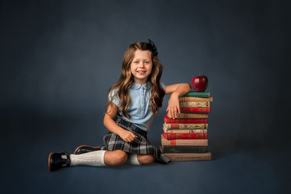 A smiling young girl in school uniform sits beside a stack of vintage books topped with a red apple on a blue background.