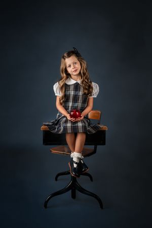 Young girl in plaid school uniform sitting on top of desk holding red apple, fine art studio portrait on dark blue background.