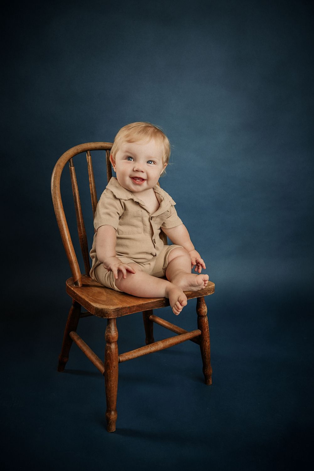 Smiling baby in beige romper sitting on a wooden Windsor chair against a dark blue studio background for fine art milestone photos in studio