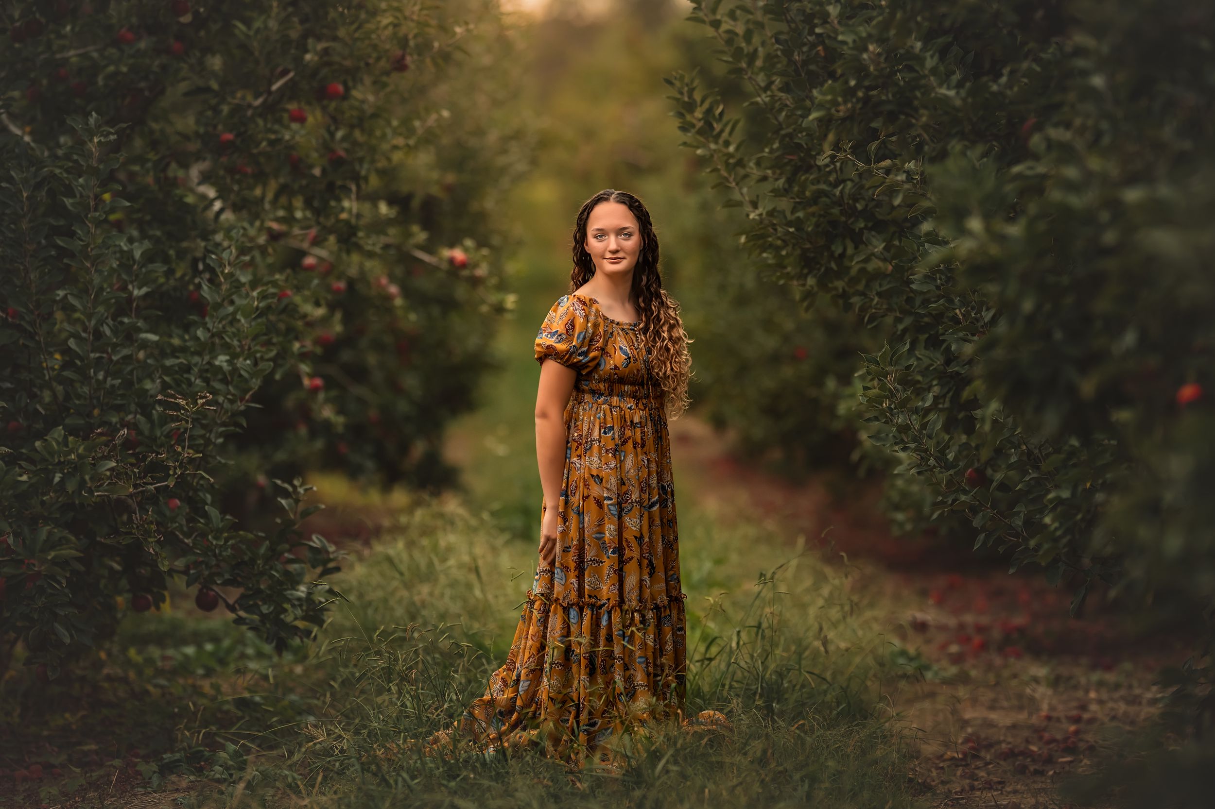 A teen girl in a gold floral dress stands among apple trees in a sunlit orchard during golden hour for fine art portrait session.