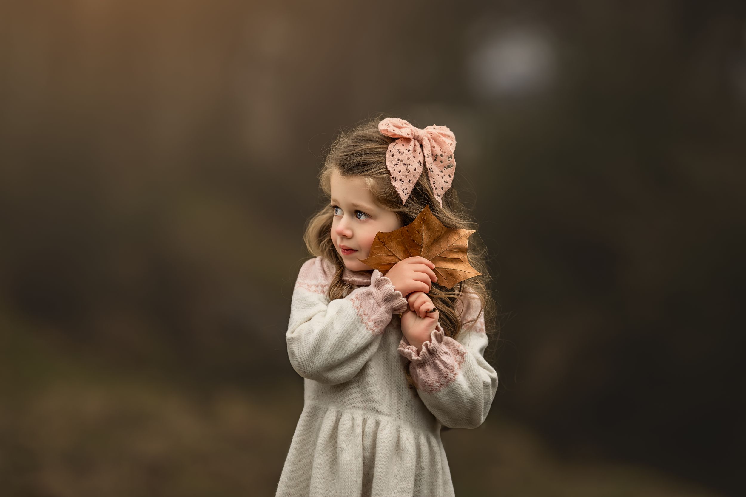 A child in a grey dress wearing a pink bow stands outdoors in soft lighting while hugging themselves.