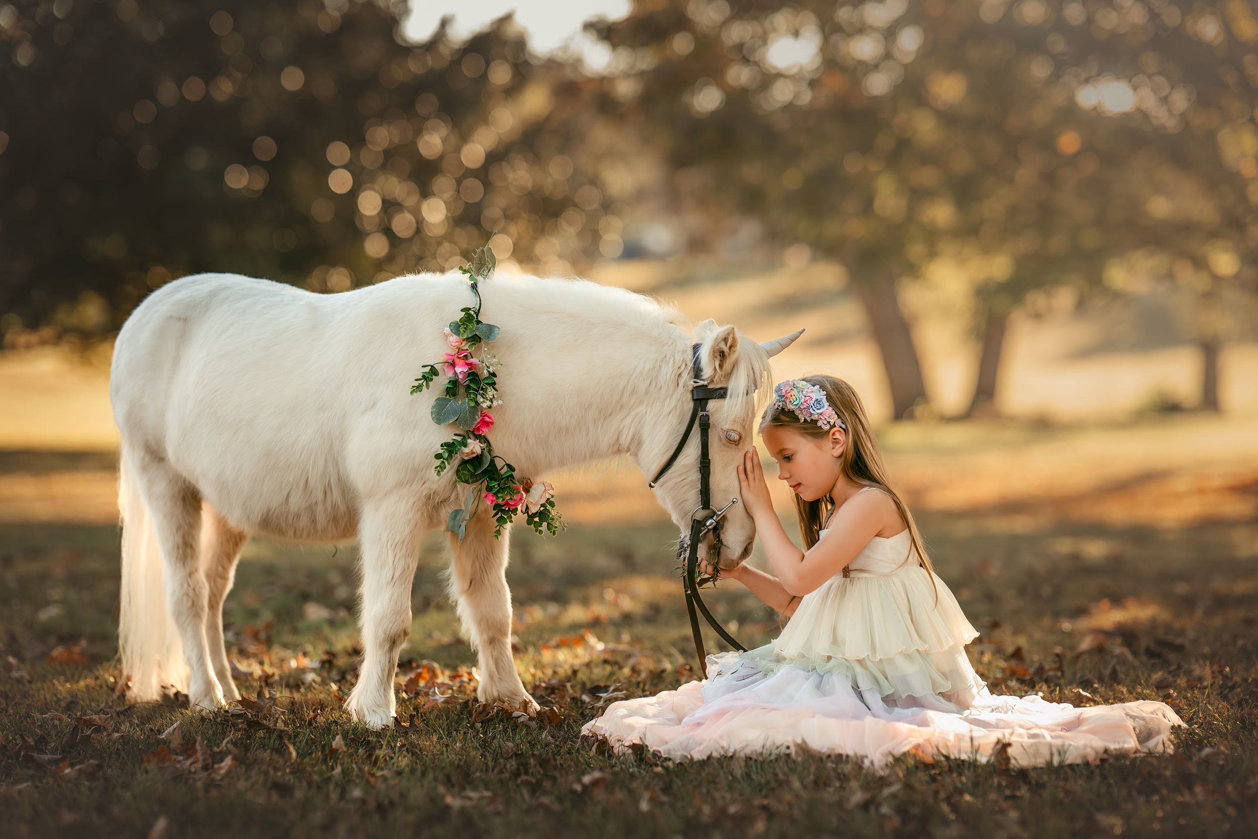 Young girl in pastel dress sits on the ground and gently touches the nose of a white unicorn pony with a floral wreath.