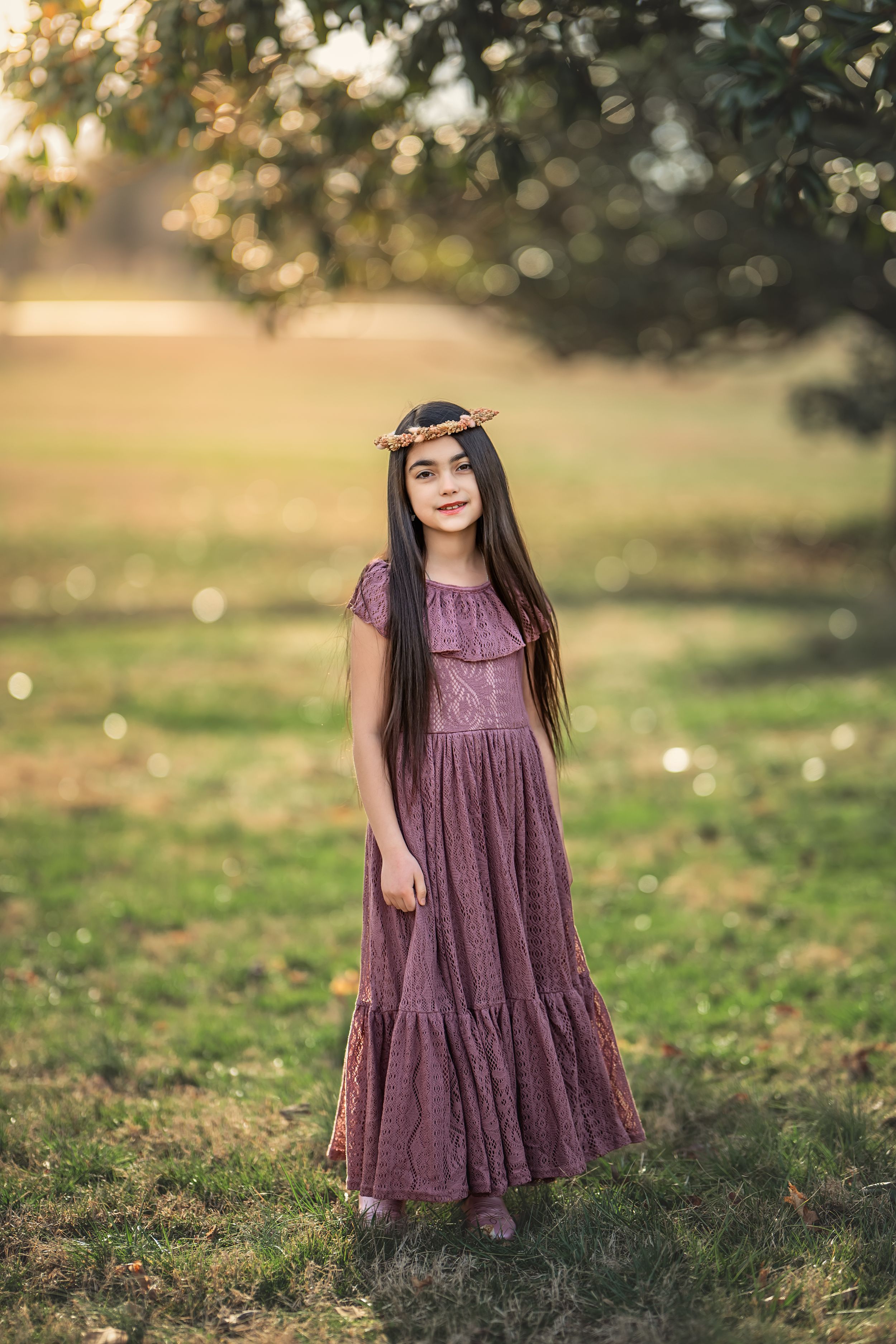 A young woman in a flowy mauve dress and floral crown stands in a sunlit garden setting.