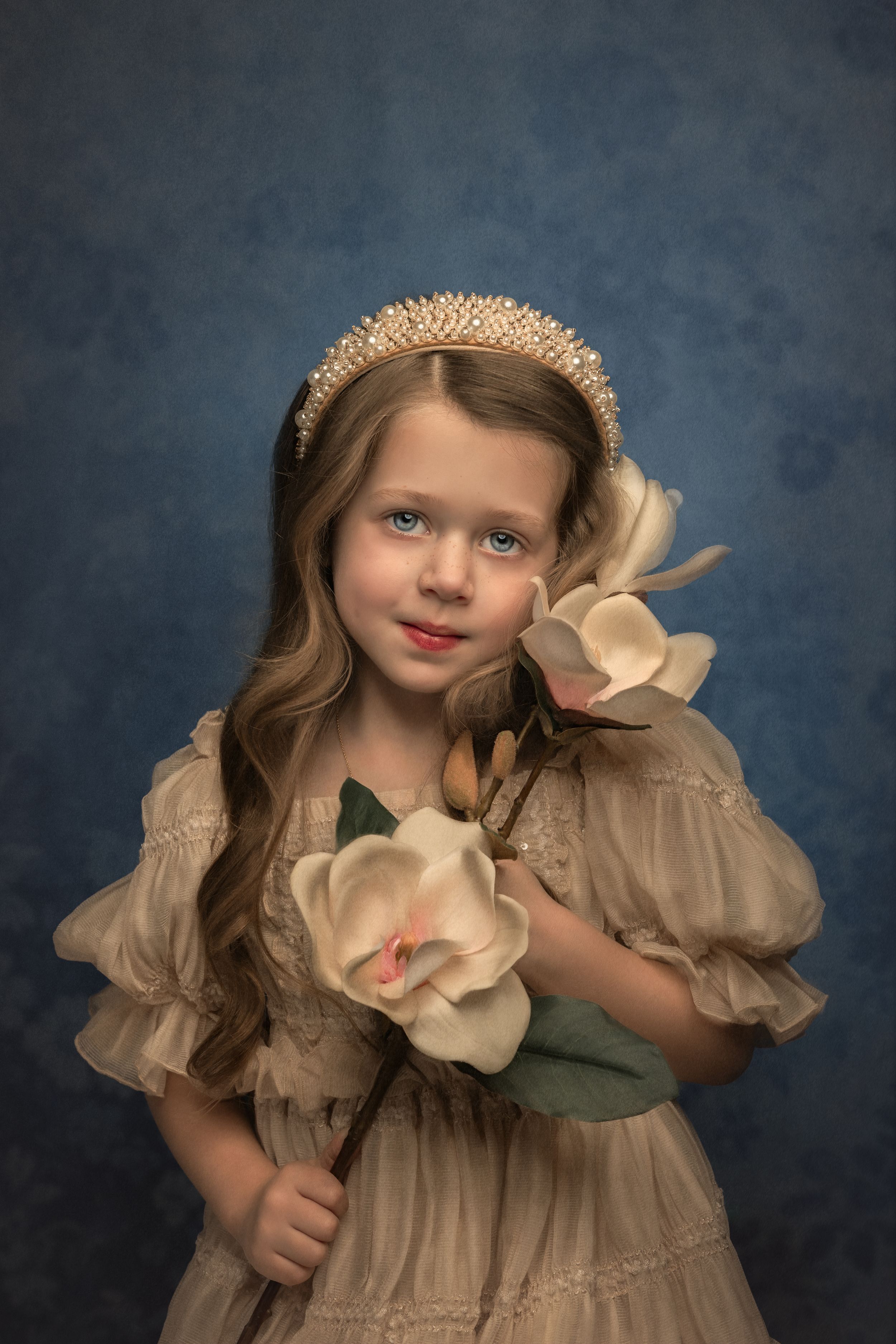Fine art portrait of a young child in vintage cream dress holding a large magnolia flower against dark blue backdrop.