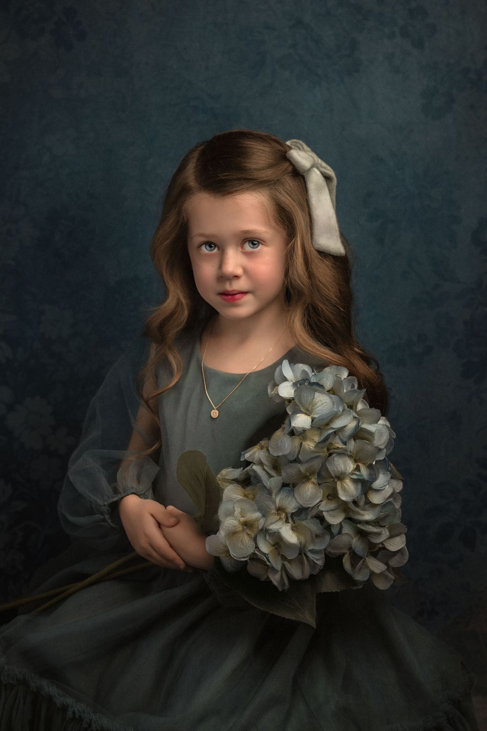 Moody fine art portrait of a child in a blue dress holding hydrangea flowers against textured dark blue backdrop.