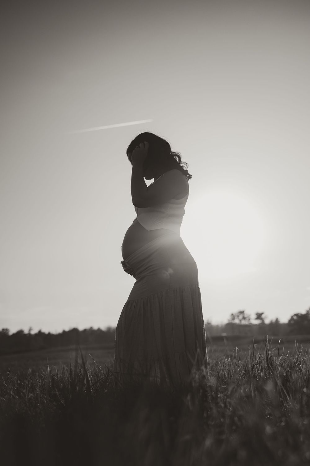 Black and white silhouette of a pregnant woman standing in a field at sunset, cradling her baby bump.