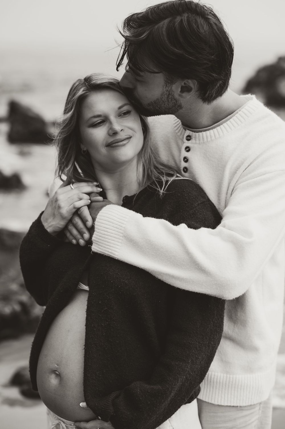 Black and white maternity photo of a couple embracing on a beach, man kissing pregnant woman's cheek tenderly.