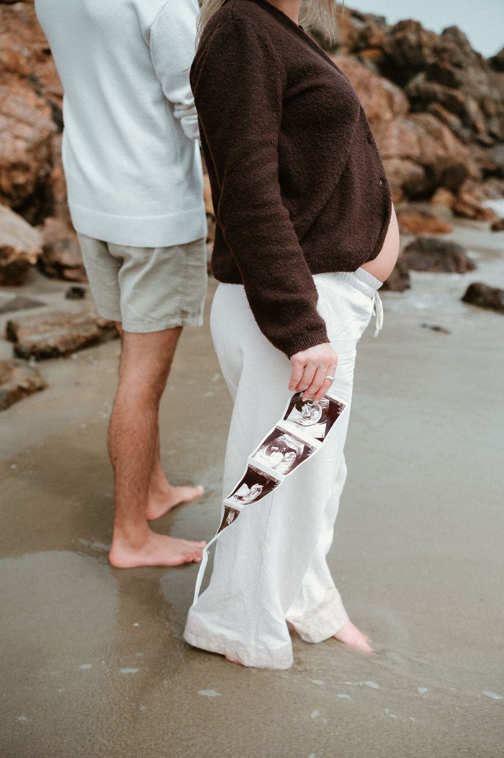Pregnant couple standing barefoot on beach, woman in white pants holding ultrasound photos, man in grey shorts beside her.
