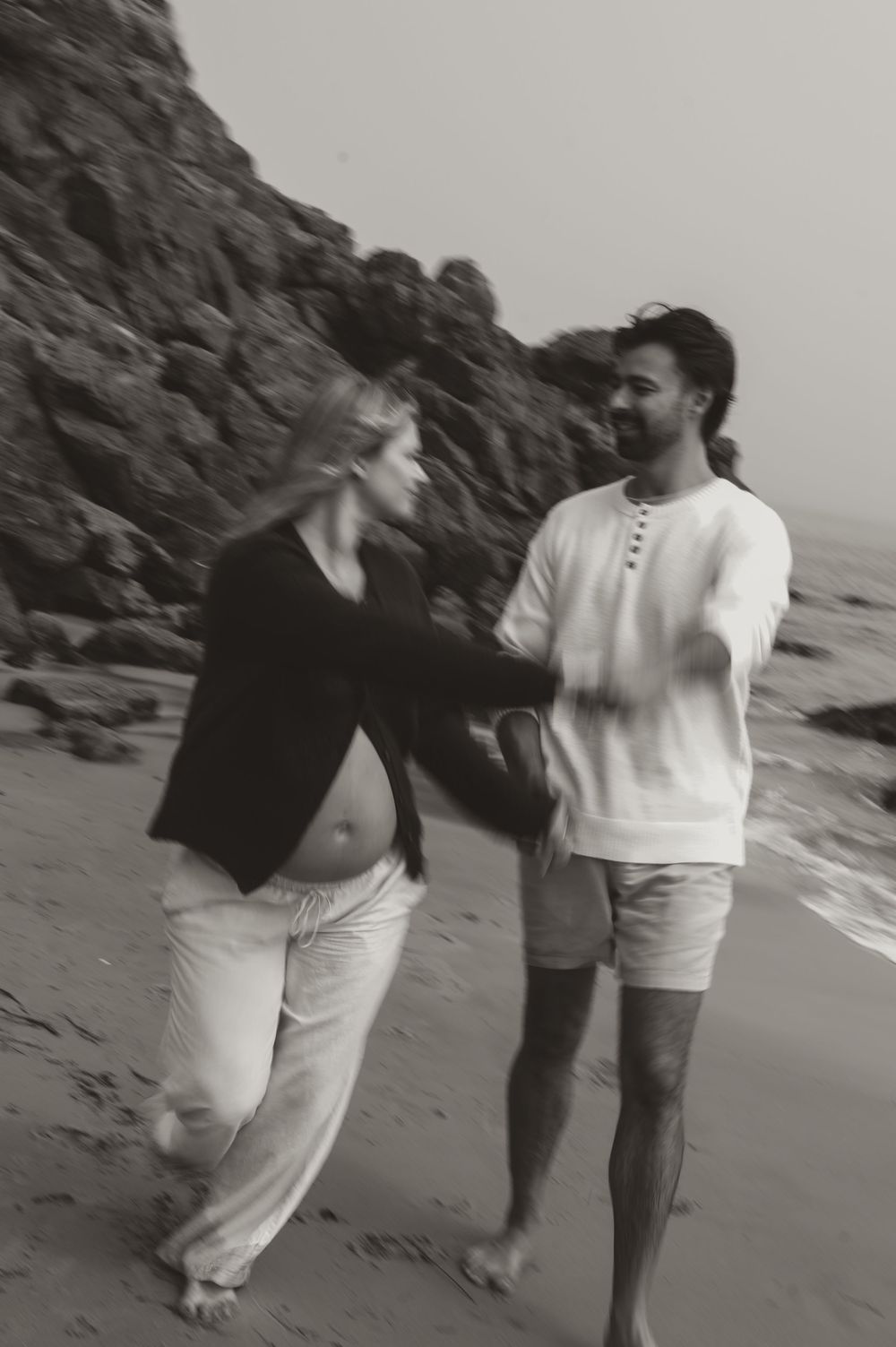 Black and white photo of a pregnant couple walking hand in hand on a rocky beach, sharing a tender moment.