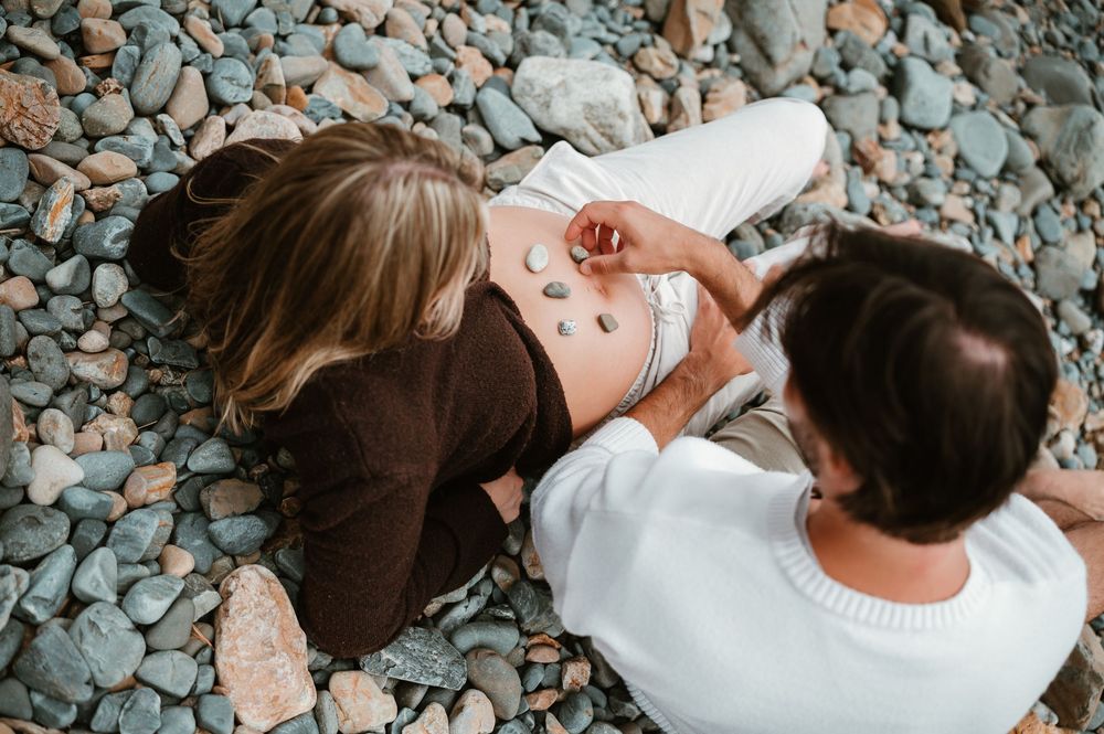Couple sitting on rocky beach, arranging small stones on pregnant woman's bare belly in a tender maternity moment.