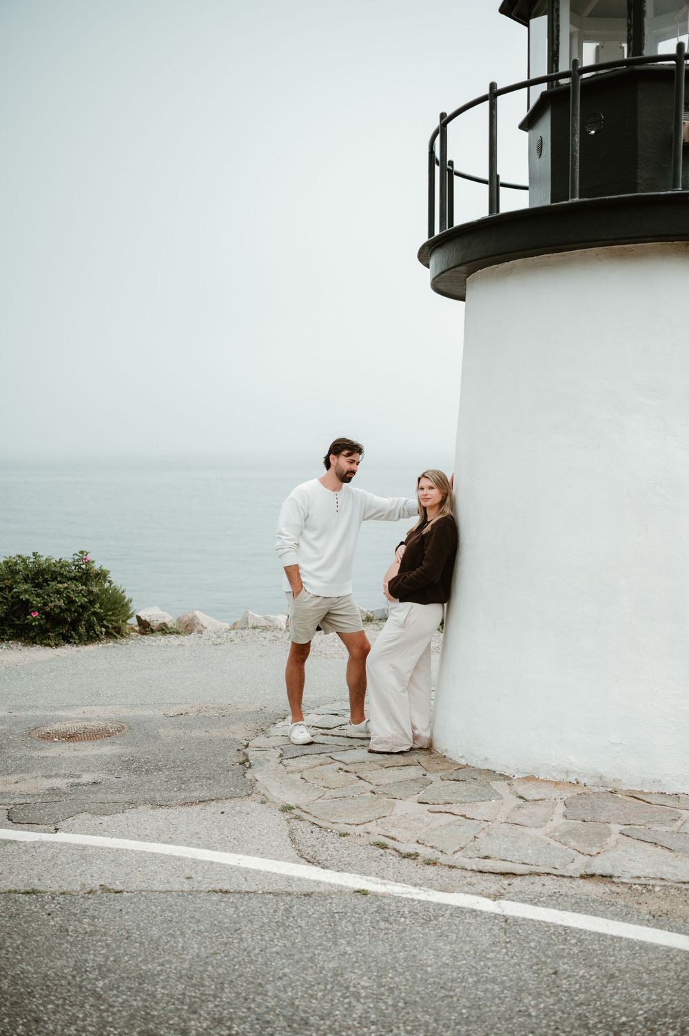 Couple posing by a white lighthouse near the ocean, man in white shirt and woman in brown top leaning against the wall.