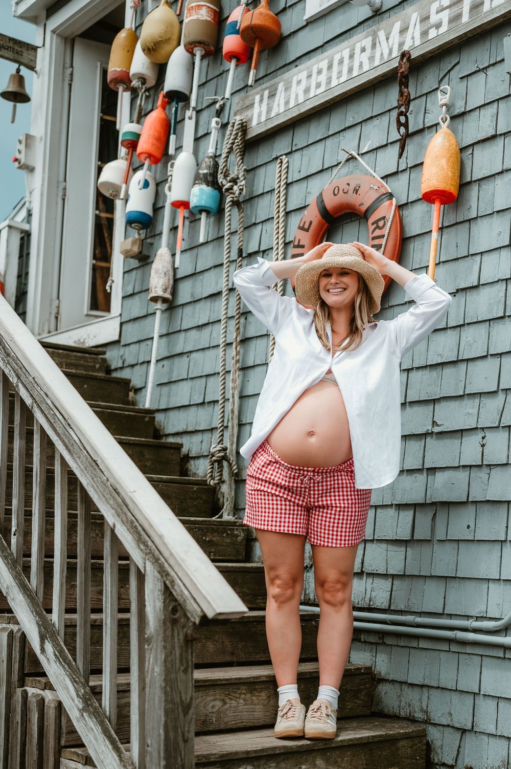 Pregnant woman in white shirt, gingham shorts, and straw hat poses on wooden steps outside a harbormaster building with colorful buoys.