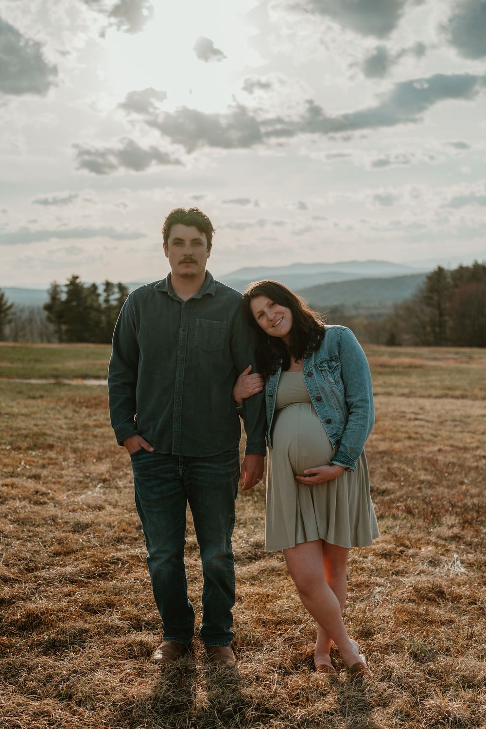 Couple posing in an open field at sunset, pregnant woman in denim jacket holding baby bump, mountains in background.